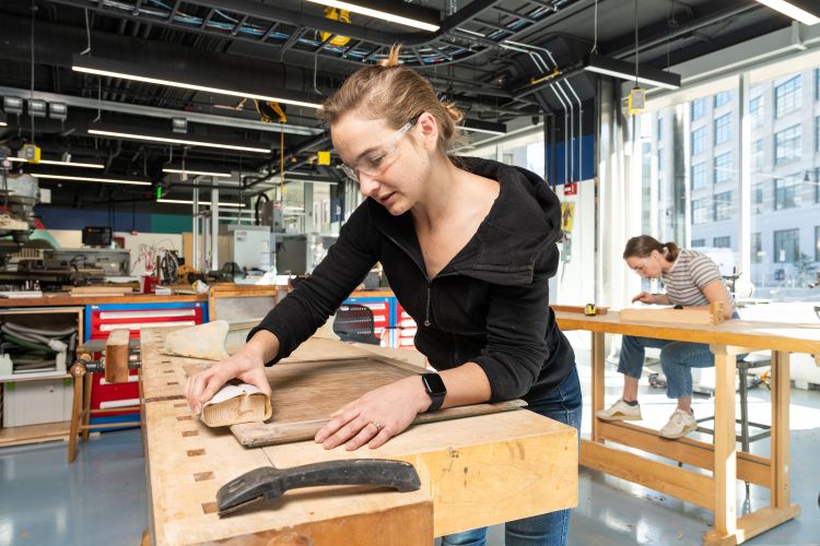 Tess Smidt leans over a butcherblock workbench sanding a board. She is in a large workroom with tall windows, high ceilings showing the mechanicals, and a clutter of equipment. Another woman can be seen at right, also leaning over a butcherblock workbench. and there are 