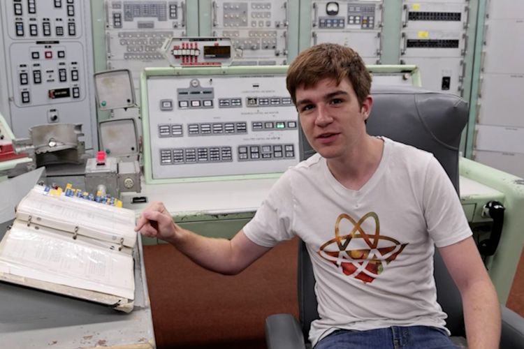 James Dingley sits in an old control room. He is wearing a white T shirt featuring the atom symbol, and he is pointing at a thick open 3-ring notebook. Behind him are an array of machines.