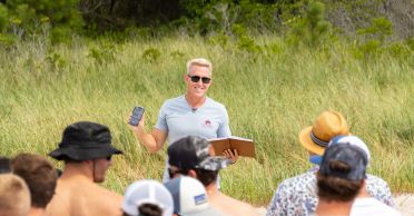 Chris Schell stands on a beach with tall grasses behind him holding a notebook and a mobile device addressing a crowd of people