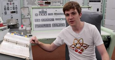 James Dingley sits in an old control room. He is wearing a white T shirt featuring the atom symbol, and he is pointing at a thick open 3-ring notebook. Behind him are an array of machines.