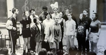 An old black and white photo shows a group of singers in front of a blackboard on which the word Chorallaries is written. The singers are wearing an assortment of garments, including a feather headress, a bandana, and a bowtie. The floor is littered with trash. The heads of some audience members appear in the foreground.