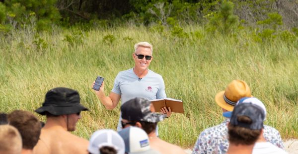 Chris Schell stands on a beach with tall grasses behind him holding a notebook and a mobile device addressing a crowd of people