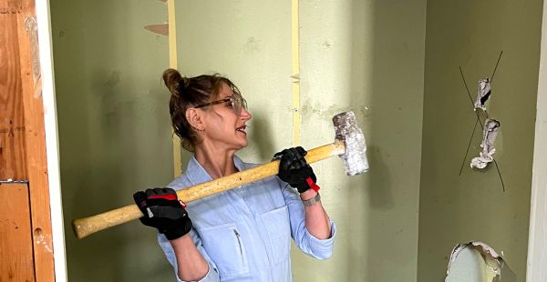 Jessica Banks wields a sledgehammer in position to strike. She is wearing gloves and coveralls and looking toward a wall with large holes in it.